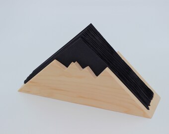 May include: A wooden napkin holder with a mountain-like design, holding a stack of black napkins. The holder is made of light-colored wood and has a simple, modern aesthetic. The napkins are neatly stacked, creating a contrast with the wood.