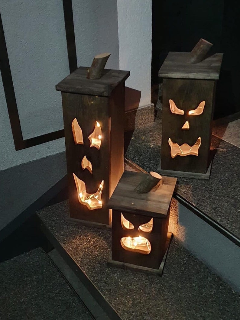May include: Three wooden Halloween lanterns with carved jack-o'-lantern faces. Each lantern has a wooden top with a small branch attached. The lanterns are lit from within, creating a spooky glow. The lanterns are different sizes.