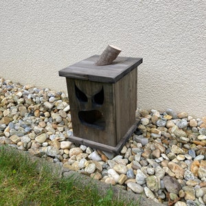 May include: A wooden birdhouse with a spooky face carved into the front. The birdhouse is stained a dark gray color and has a small wooden branch protruding from the top. It sits on a bed of rocks and grass.