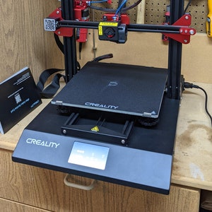 K&ouml;nnte beinhalten: Ein schwarzer 3D-Drucker mit rotem Rahmen und einem schwarz-wei&szlig;en Display. Der Drucker steht auf einer Holzoberfl&auml;che und tr&auml;gt den Schriftzug "CREALITY" auf der Vorderseite.