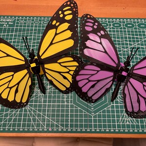 K&ouml;nnte beinhalten: Zwei 3D-gedruckte Schmetterlinge, einer gelb und einer lila, werden auf einer gr&uuml;nen Schneidematte pr&auml;sentiert. Die Schmetterlinge haben schwarze Umrisse und komplizierte Fl&uuml;gelmuster.
