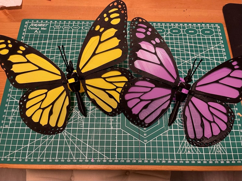 K&ouml;nnte beinhalten: Zwei 3D-gedruckte Schmetterlinge, einer gelb und einer lila, werden auf einer gr&uuml;nen Schneidematte pr&auml;sentiert. Die Schmetterlinge haben schwarze Umrisse und komplizierte Fl&uuml;gelmuster.
