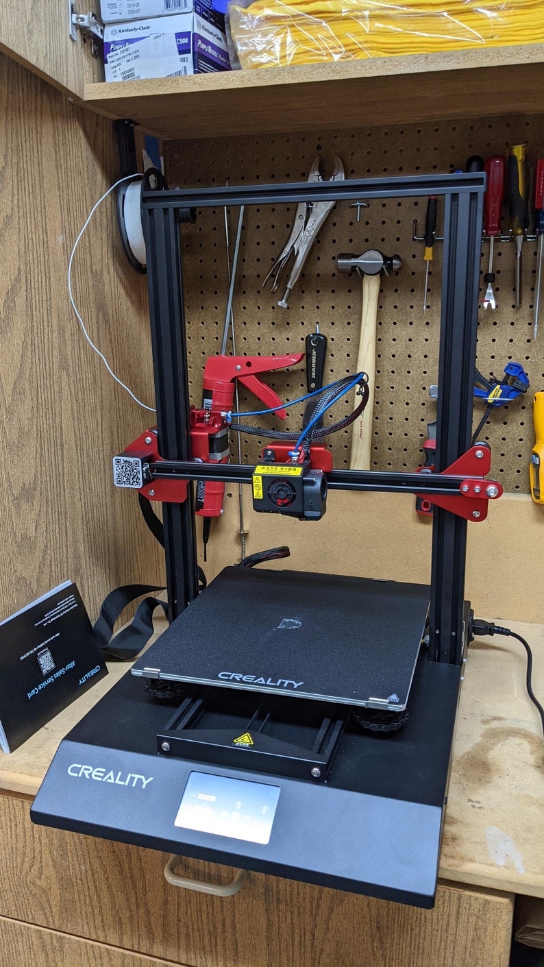 K&ouml;nnte beinhalten: Ein schwarzer 3D-Drucker mit rotem Rahmen und einem schwarz-wei&szlig;en Display. Der Drucker steht auf einer Holzoberfl&auml;che und tr&auml;gt den Schriftzug "CREALITY" auf der Vorderseite.