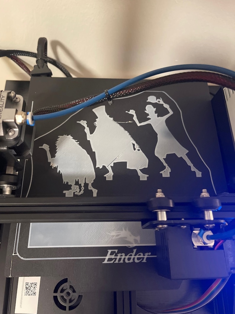 K&ouml;nnte beinhalten: Ein 3D-gedrucktes Modell von drei Figuren, einem Geist, einer Frau und einer Kreatur, auf einem schwarzen 3D-Druckbett. Das Modell ist in wei&szlig;em Filament gedruckt. Der Drucker ist mit "Ender" beschriftet.