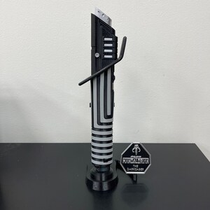 K&ouml;nnte beinhalten: Eine schwarze und silberne Lichtschwert-Replik mit einem St&auml;nder und einem Metallabzeichen mit der Aufschrift "STAR WARS THE MANDALORIAN THE DARKSABER".