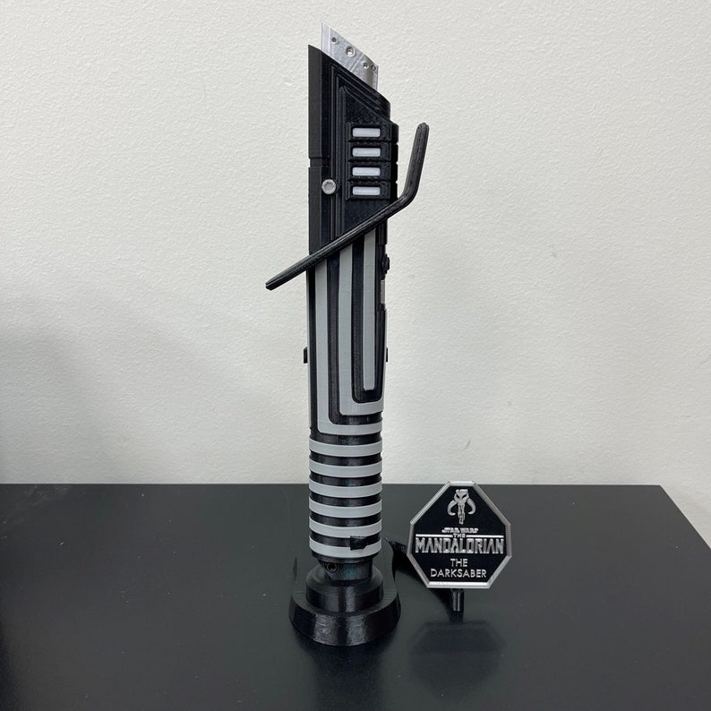 K&ouml;nnte beinhalten: Eine schwarze und silberne Lichtschwert-Replik mit einem St&auml;nder und einem Metallabzeichen mit der Aufschrift "STAR WARS THE MANDALORIAN THE DARKSABER".