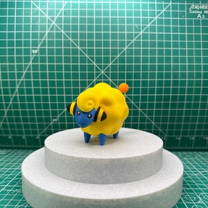 May include: A yellow and blue sheep figurine with black markings on its head and a small orange tail. The figurine is standing on a white two-tiered cake stand.