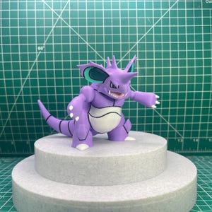 Puede incluir: Una figura morada y blanca de una criatura con una larga cola, cuernos y orejas grandes. La figura está sobre una plataforma circular gris de dos niveles. El fondo es una esterilla de corte verde.