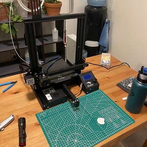 K&ouml;nnte beinhalten: Ein schwarzer 3D-Drucker mit einer gr&uuml;nen Schneidematte auf einem Holztisch. Der Drucker hat eine wei&szlig;e Filamentspule und ein blaues und schwarzes Kabel. Die Schneidematte hat einen wei&szlig;en Kreis in der Mitte.