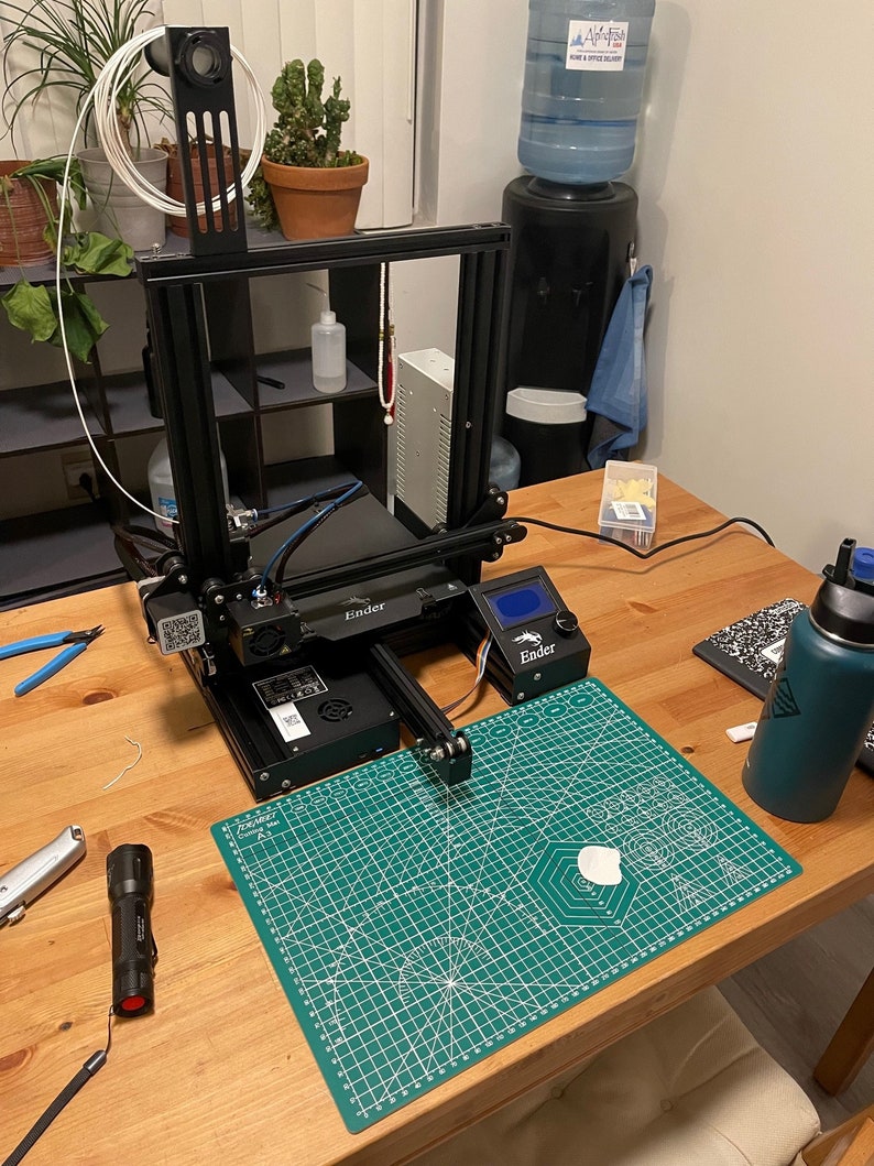 K&ouml;nnte beinhalten: Ein schwarzer 3D-Drucker mit einer gr&uuml;nen Schneidematte auf einem Holztisch. Der Drucker hat eine wei&szlig;e Filamentspule und ein blaues und schwarzes Kabel. Die Schneidematte hat einen wei&szlig;en Kreis in der Mitte.