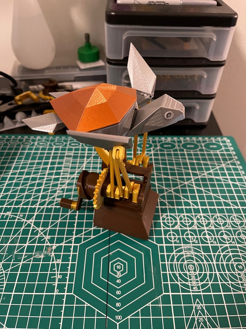 K&ouml;nnte beinhalten: Eine 3D-gedruckte Steampunk-Schildkr&ouml;te mit brauner Schale, silbernen Beinen und goldenen Zahnr&auml;dern. Die Schildkr&ouml;te ist auf einem Holzsockel mit einer Kurbel montiert.