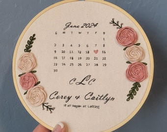 May include: Embroidered hoop art with a calendar for June 2024, featuring a heart on the 13th and the text "Corey & Caitlyn It all began at Lotties!" with floral embellishments.