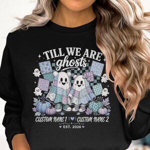 Peut inclure: Un sweat-shirt noir avec un motif incluant le texte TILL WE ARE ghosts, deux personnages fantômes vêtus de draps en patchwork, des citrouilles et de petits fantômes. Sous les personnages, le texte indique CUSTOM NAME 1, un cœur, CUSTOM NAME 2, et EST. 2026.