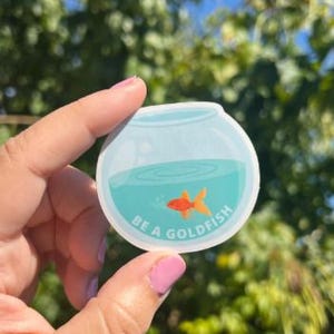 May include: A round sticker featuring a cartoon goldfish in a bowl. The bowl has blue water and the text "BE A GOLDFISH" in white. The goldfish is orange. The sticker is held against a background of green foliage and blue sky.