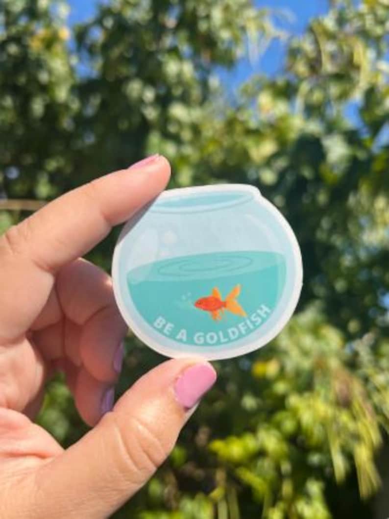 May include: A round sticker featuring a cartoon goldfish in a bowl. The bowl has blue water and the text "BE A GOLDFISH" in white. The goldfish is orange. The sticker is held against a background of green foliage and blue sky.