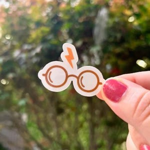 May include: A white sticker featuring a pair of round glasses with brown frames and a lightning bolt in orange. The sticker is held up against a blurred green background.