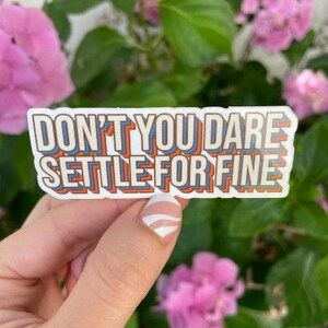 May include: A sticker with the text "Don't you dare settle for fine" in a retro, multi-colored font. The sticker is held in a hand with white nail polish.