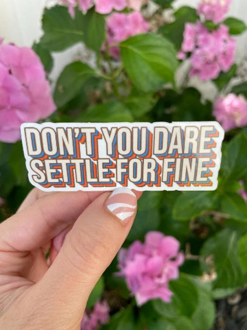 May include: A sticker with the text "Don't you dare settle for fine" in a retro, multi-colored font. The sticker is held in a hand with white nail polish.