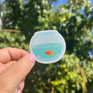 May include: A round sticker featuring a cartoon goldfish in a bowl with teal water. The sticker has the text "BE A GOLDFISH" in white. The background is a blurred view of green foliage and a blue sky.
