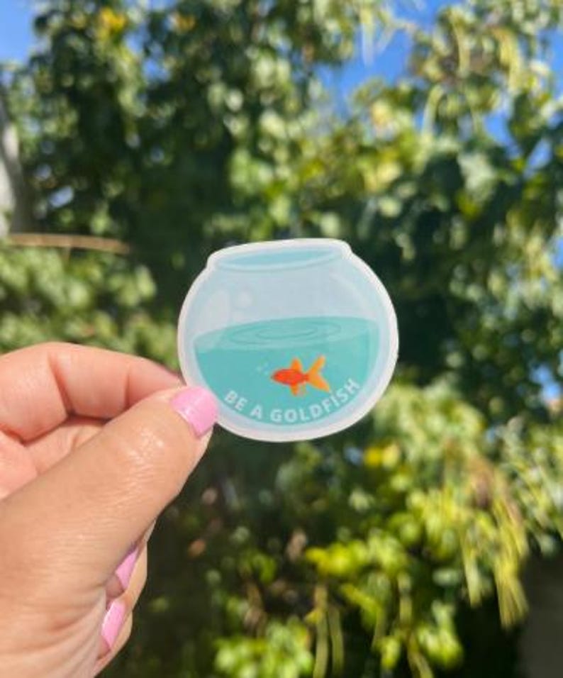 May include: A round sticker featuring a cartoon goldfish in a bowl with teal water. The sticker has the text "BE A GOLDFISH" in white. The background is a blurred view of green foliage and a blue sky.