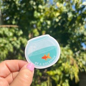 May include: A round sticker featuring a goldfish in a bowl. The bowl is light blue with a teal interior and a small orange goldfish. The sticker has the text "BE A GOLDFISH" in white, curved around the bowl.