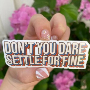 May include: White sticker with the text "Don't you dare settle for fine" in a retro, colorful font. The sticker is held by a hand with white flowers on the nails.