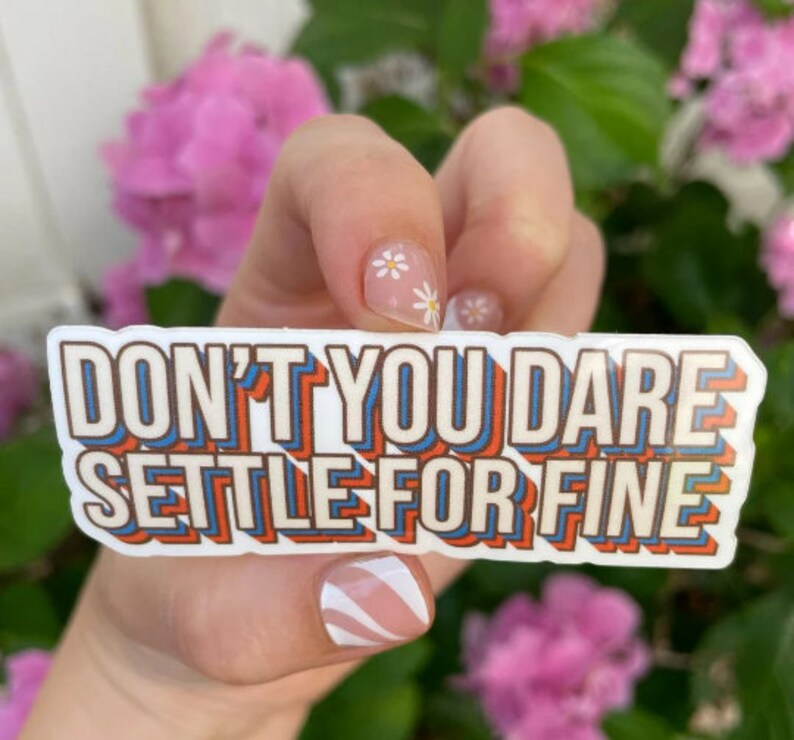 May include: White sticker with the text "Don't you dare settle for fine" in a retro, colorful font. The sticker is held by a hand with white flowers on the nails.