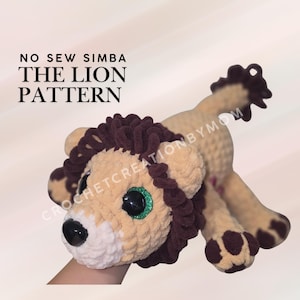 May include: A crocheted lion plush toy with tan and white yarn, a dark brown mane, and green glittery eyes. The text NO SEW SIMBA THE LION PATTERN is visible. The lion has a black nose and dark brown paw pads on its four feet and tail tip.