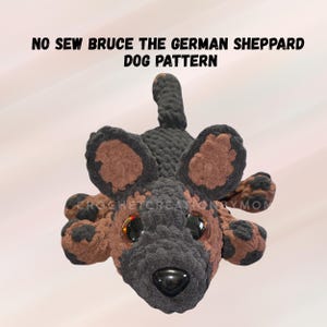 May include: A plush German Shepherd dog toy made of dark brown and black yarn with large, glossy black safety eyes and a black plastic nose. The text NO SEW BRUCE THE GERMAN SHEPPARD DOG PATTERN is displayed above the toy in black capital letters.