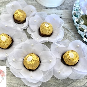 May include: White tulle flower-shaped wrappers with pearl accents, each holding a golden chocolate candy. The wrappers are arranged on a clear glass platter.