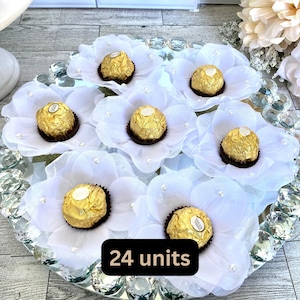 以下が含まれることがあります： 金色のホイルで包まれた7つの丸いチョコレートが、小さな白いビーズで飾られた白い布製の花の中にそれぞれ配置されています。黒い長方形のラベルには「24 units」というテキストが表示されています。