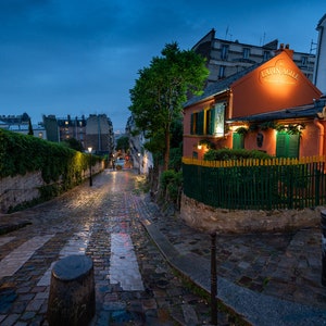 May include: A cobblestone street in Paris, France, at dusk. The street is wet from rain and leads up to a building with a sign that reads "Au Lapin Agile Cabaret".