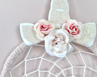 May include: White dream catcher with a white unicorn horn and pink flowers on top. The dream catcher is made of white yarn and has a white feather trim.