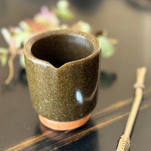 Può includere: Una piccola tazza in ceramica verde oliva macchiettata con un beccuccio e una base marrone chiaro. Una cannuccia di metallo con un'estremità a motivi è appoggiata accanto alla tazza. La tazza è probabilmente per bere tè o una bevanda simile.