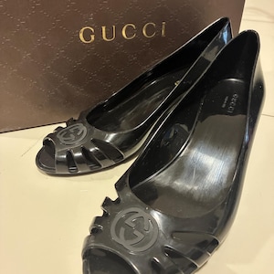 Puede incluir: Un par de zapatos de tacón peep-toe de gelatina negra con un diseño recortado en la parte delantera y un emblema circular con el logotipo de Gucci. Los zapatos están colocados frente a una caja marrón de la marca Gucci con un patrón repetitivo.