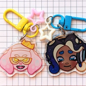 May include: Two colorful keychains featuring cartoon characters. One keychain is pink and white with a character wearing sunglasses and a crown. The other keychain is blue and white with a character wearing headphones and a crown.