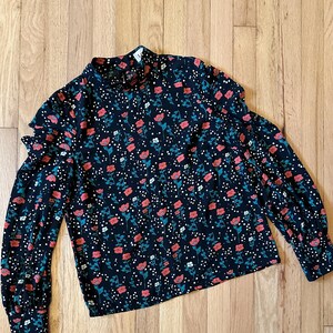 May include: A black long-sleeved blouse with a floral print featuring red, white, and green flowers on a black background. The blouse has a high neck and ruffled sleeves.