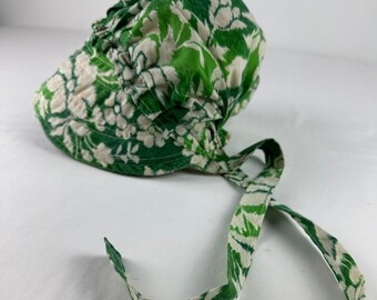 Puede incluir: Un gorro con estampado floral verde y blanco con cintas. El gorro está hecho de una tela suave y presenta un diseño fruncido alrededor de la cabeza. Las cintas también tienen el mismo diseño floral.