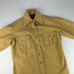 Może przedstawiać: Koszula w kolorze beżowym zapinana na guziki, z dwoma dużymi kieszeniami na klapki na piersi. Koszula ma podszewkę pikowaną i metkę z napisem "Woolrich, Woolrich, Inc., New York, N.Y."