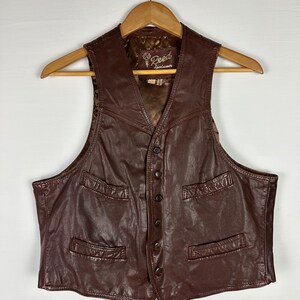 May include: A brown leather vest with a button closure and multiple pockets. The vest has a label that reads "G. Reed Leatherwear."