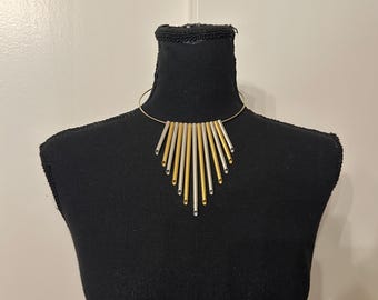 May include: A gold and silver-toned necklace with a geometric design. The necklace features a thin, circular band that sits around the neck, with vertical bars of gold and silver metal hanging down in a fan shape. The necklace is displayed on a black mannequin.