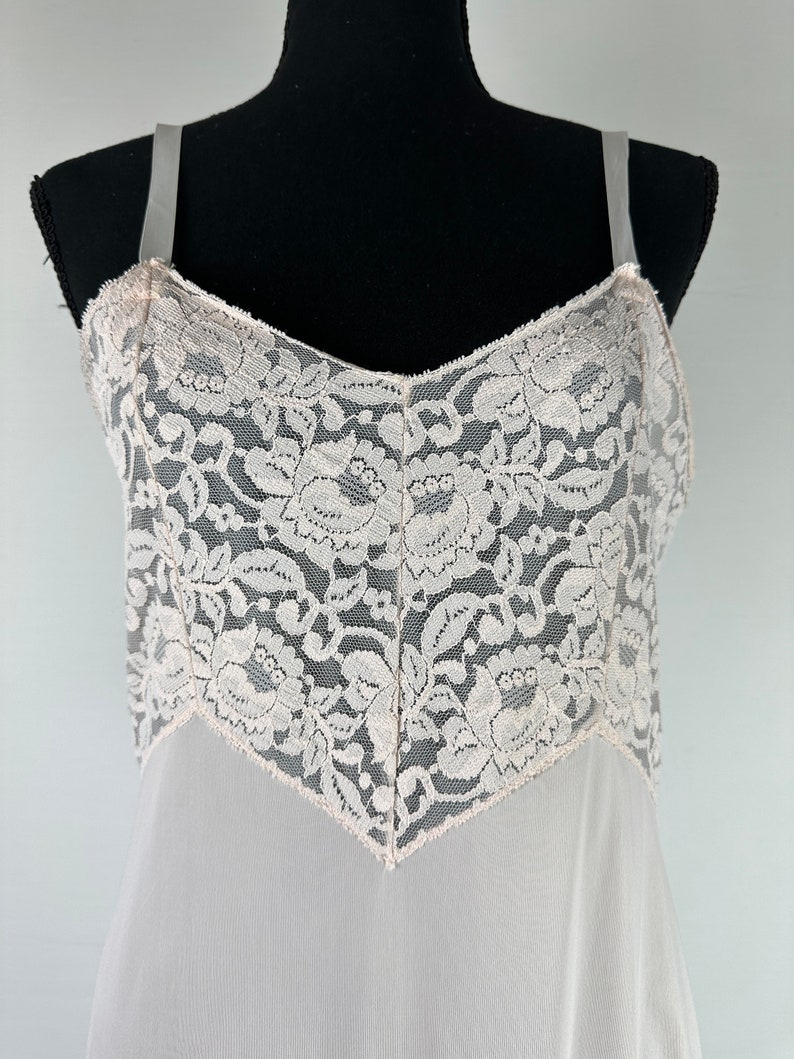 Op de afbeelding: Een witte slip van kant met delicate bloemenpatronen. De bandjes zijn dun en de halslijn is een klassieke V-hals.
