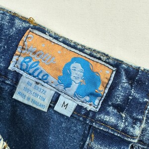 以下が含まれることがあります： ゴールドとブルーのフォントで「Sassy Blue」と書かれたブルーのデニムジーンズのラベル。ラベルには、ブルーの背景に女性の顔と「RN 59372 100% COTTON MADE IN THAILAND」と「M」の文字も含まれています。