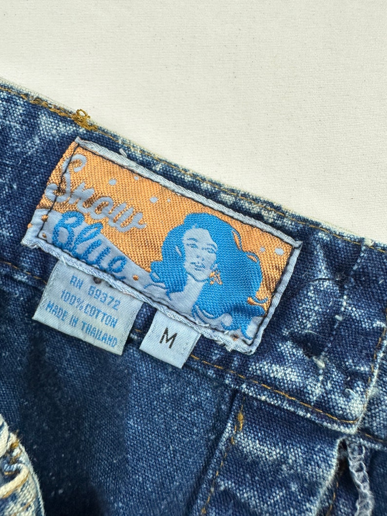 以下が含まれることがあります： ゴールドとブルーのフォントで「Sassy Blue」と書かれたブルーのデニムジーンズのラベル。ラベルには、ブルーの背景に女性の顔と「RN 59372 100% COTTON MADE IN THAILAND」と「M」の文字も含まれています。