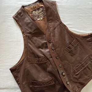 May include: A brown leather vest with a button closure and multiple pockets. The vest has a label that reads "Reed Sportswear First in Quality".