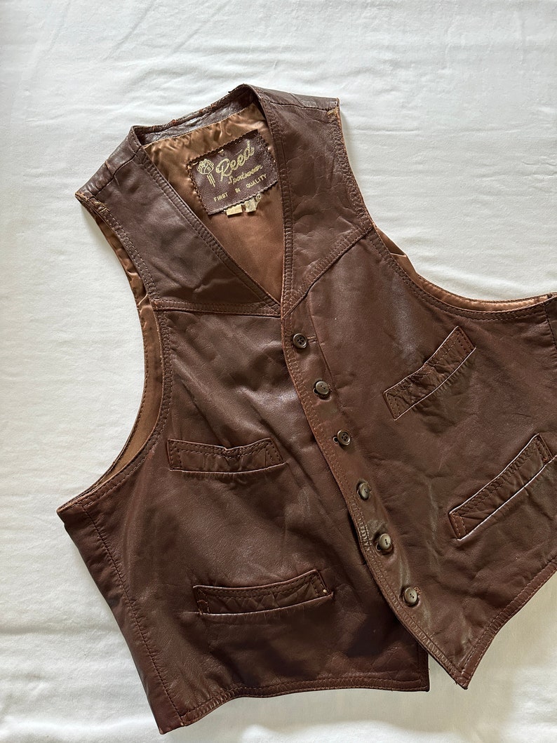May include: A brown leather vest with a button closure and multiple pockets. The vest has a label that reads "Reed Sportswear First in Quality".