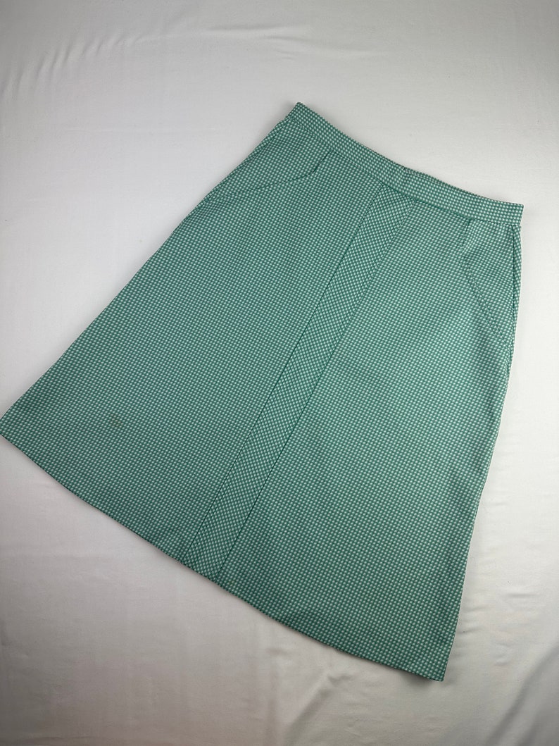 May include: A green and white gingham skirt with a straight cut and a button closure. The skirt has two pockets on the sides.
