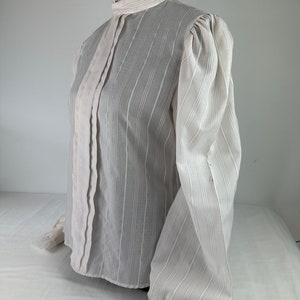 May include: A white, long-sleeved blouse with a high collar and lace trim. The blouse has a striped pattern and a slightly puffed sleeve.