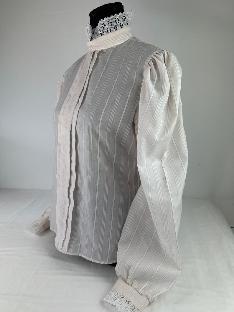 May include: A white, long-sleeved blouse with a high collar and lace trim. The blouse has a striped pattern and a slightly puffed sleeve.