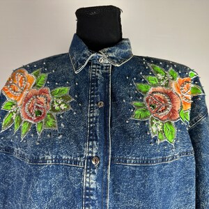 以下が含まれることがあります： オレンジ、ピンク、グリーンのバラが刺繍された、ブルーのデニムジャケット。シルバーの装飾が施されています。